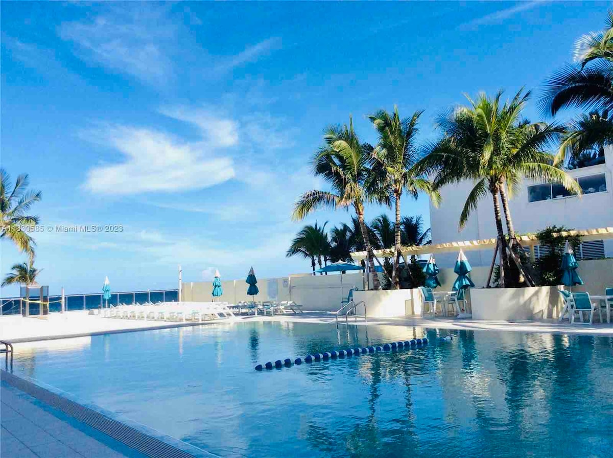 A spacious outdoor pool is surrounded by palm trees and deck chairs, providing a serene environment. The clear blue water reflects the sky, and several umbrellas offer shade. The ocean can be seen in the background, enhancing the inviting poolside atmosphere.