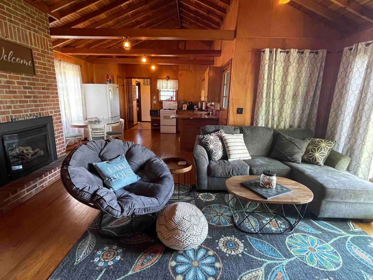 A welcoming living area features a gray sectional sofa and a round chair, arranged around a coffee table. A neutral area rug with floral patterns complements the space. Large windows allow natural light to fill the room, while a fireplace adds warmth to the setting.