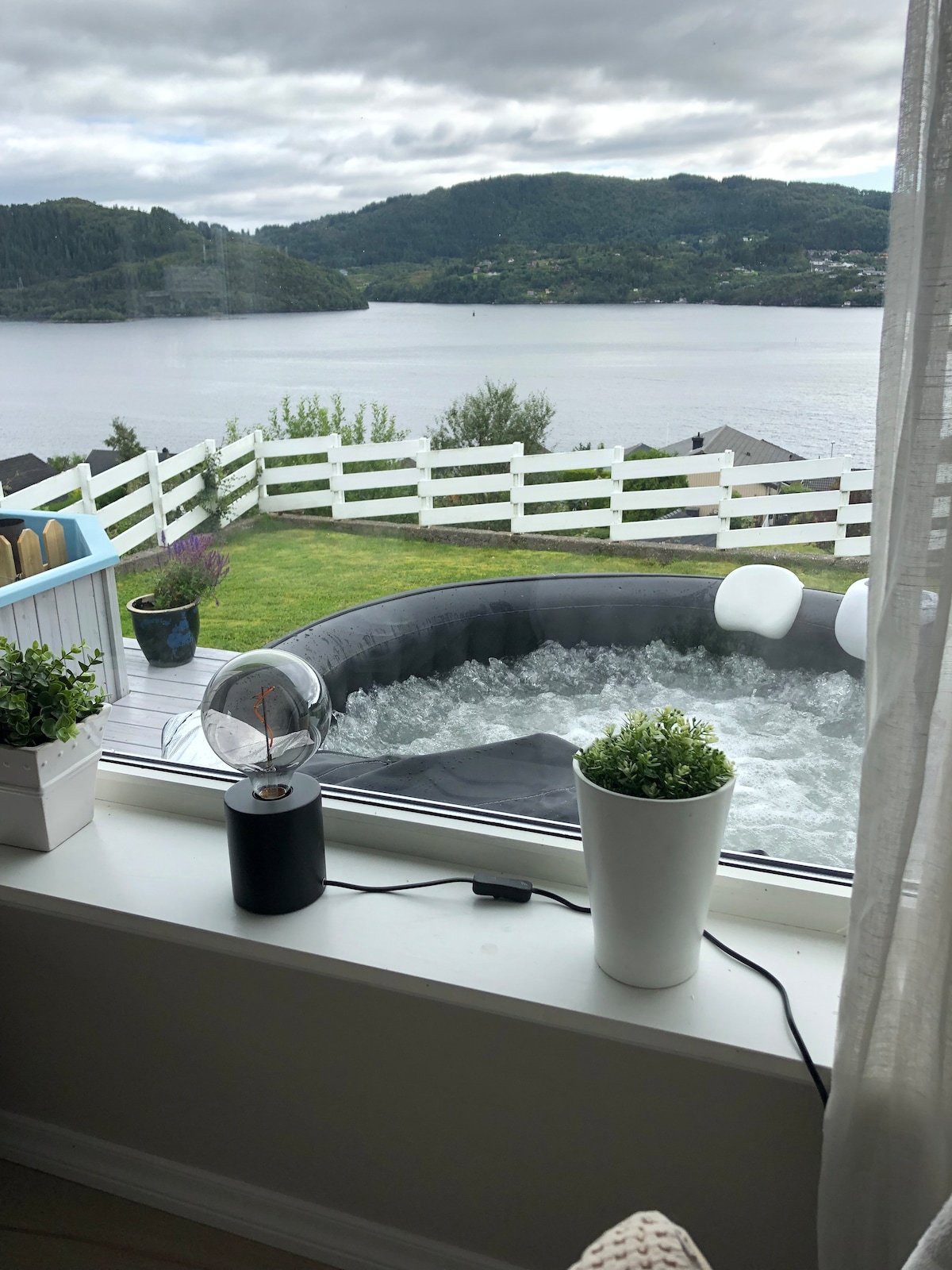 A view through a large window features a hot tub in the foreground, surrounded by greenery. Beyond the tub, a landscape of rolling hills and fjord waters is visible, complemented by a white fence outlining the property.