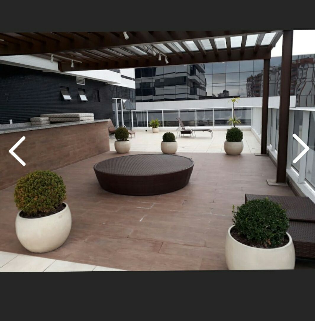 An expansive terrace area is presented, featuring a round wicker table surrounded by low hedges in decorative pots. Modern lounge chairs are positioned on one side, and a sleek railing provides a view of the surrounding buildings.