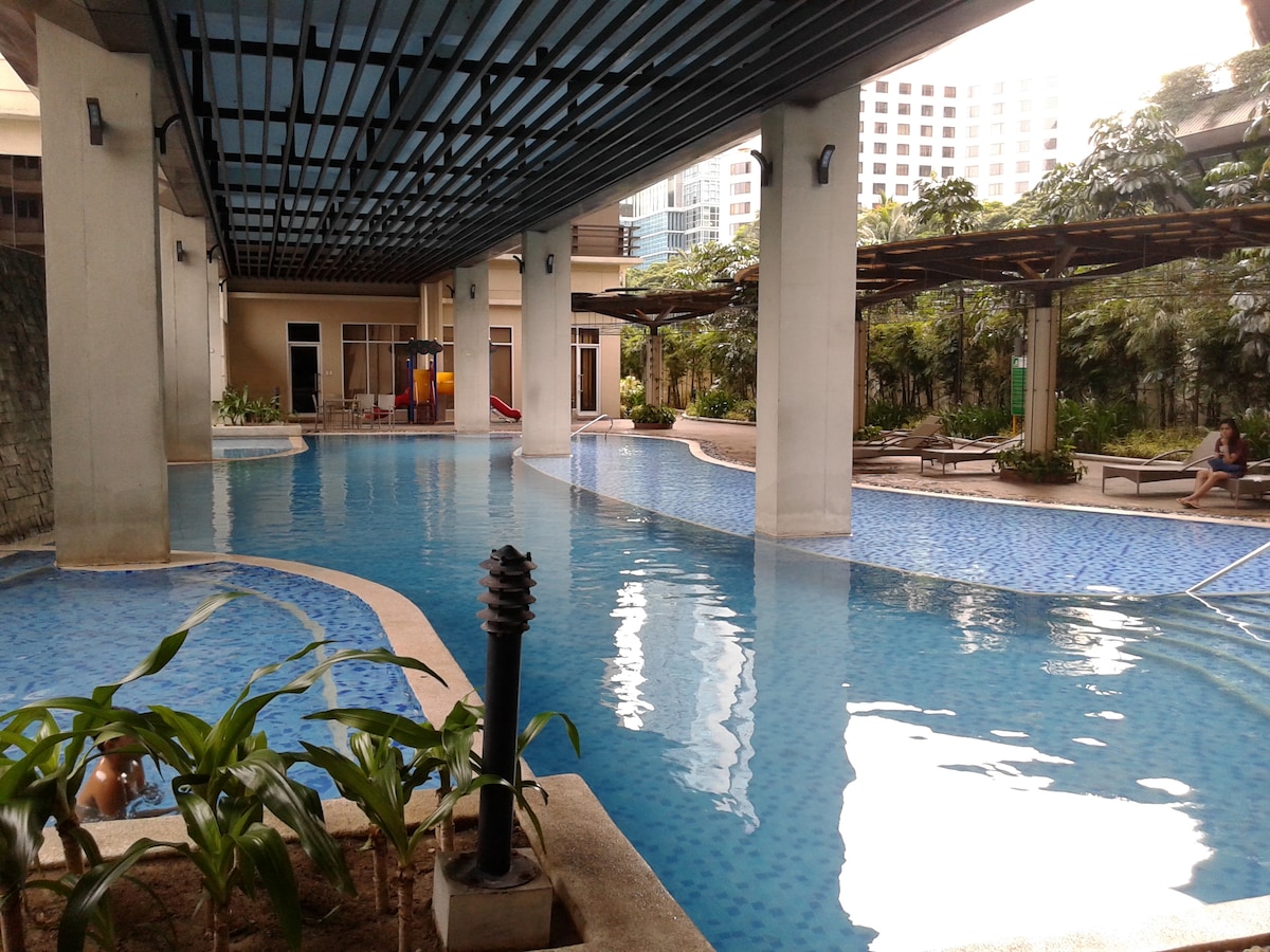 A resort-style pool is surrounded by a landscaped area featuring greenery and lounge chairs. The water reflects the overhanging structure, enhancing the tranquil atmosphere. Sunlight filters through the openings, creating a serene environment perfect for relaxation.