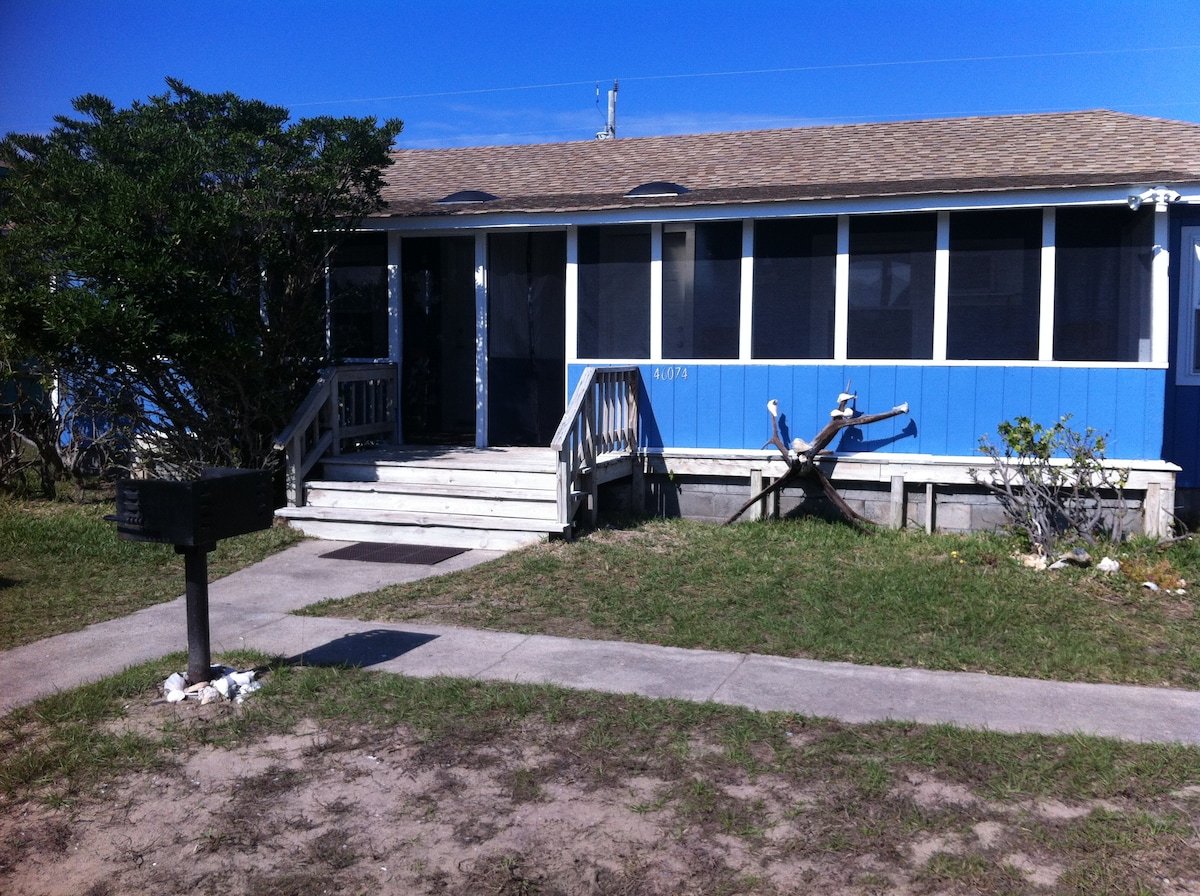 A quaint beach cottage exterior features blue wooden siding and a slanted roof under a clear sky. The entrance is accessible by a wooden ramp leading to a covered porch with screening. A path lined with grass and small rocks leads up to the front of the home.