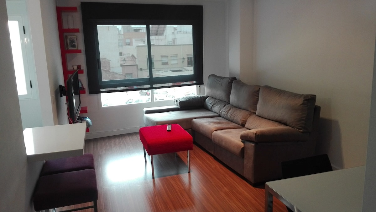 A modern living area features a large corner sofa and a red accent table. Natural light fills the space through a wide window, highlighting the warm wood flooring. A small dining table is visible, complemented by two purple stools against a white wall.