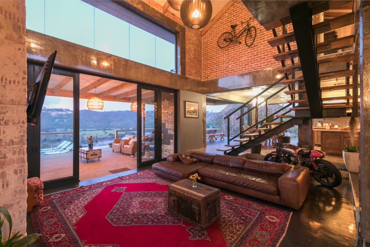 A spacious living area features a comfortable leather sofa arranged on a decorative rug, with large glass doors providing views of the outdoor patio and surrounding landscape. A staircase leads to the upper level, and bicycles are displayed decoratively on the wall.