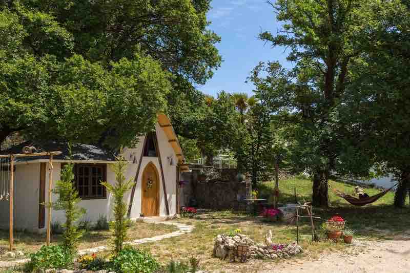 The exterior of a charming cottage is revealed, featuring an arched door under a sloping roof surrounded by lush greenery. A pathway leads to the entrance, while colorful flower pots enhance the inviting atmosphere. A shaded hammock is visible nearby, adding to the tranquil setting.