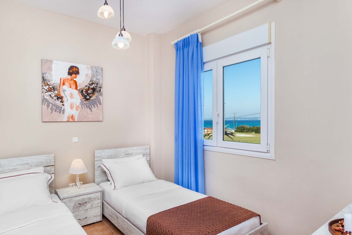 A light-filled bedroom features two single beds, each with crisp white linens and a decorative brown throw. A small bedside table with a lamp is positioned between the beds. A large window offers a view of the outdoors, framed by soft blue curtains.