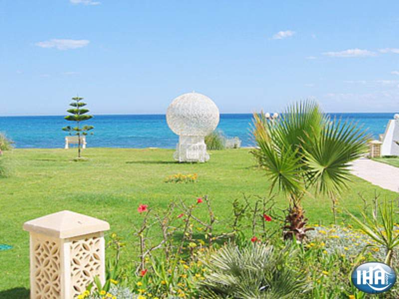 A well-maintained garden area is visible, featuring manicured green grass and various tropical plants. A decorative white structure resembling a globe is positioned prominently. The sea can be seen in the background, with a clear blue sky overhead, enhancing the tranquil coastal environment.
