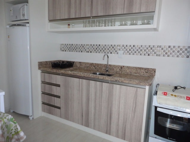 A modern kitchen space is presented with light wood cabinetry and a sleek granite countertop. Glass shelves display dishware, and a stainless steel sink is positioned beneath the window. A white refrigerator and stove complete the kitchen arrangement, contributing to a functional cooking area.