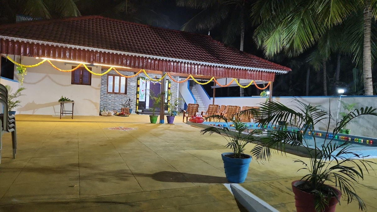 A spacious outdoor area is featured, showcasing a well-lit swimming pool surrounded by tropical greenery. The building has a traditional design with a red-tiled roof and festive decorations. Comfortable seating arrangements are visible, making it suitable for gatherings and celebrations.