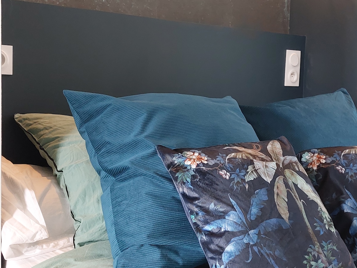 A cozy sleeping area is shown with a variety of soft pillows in teal and floral patterns. The headboard is dark, complementing the textured bedding. A natural light source is visible, enhancing the inviting feel of the space.