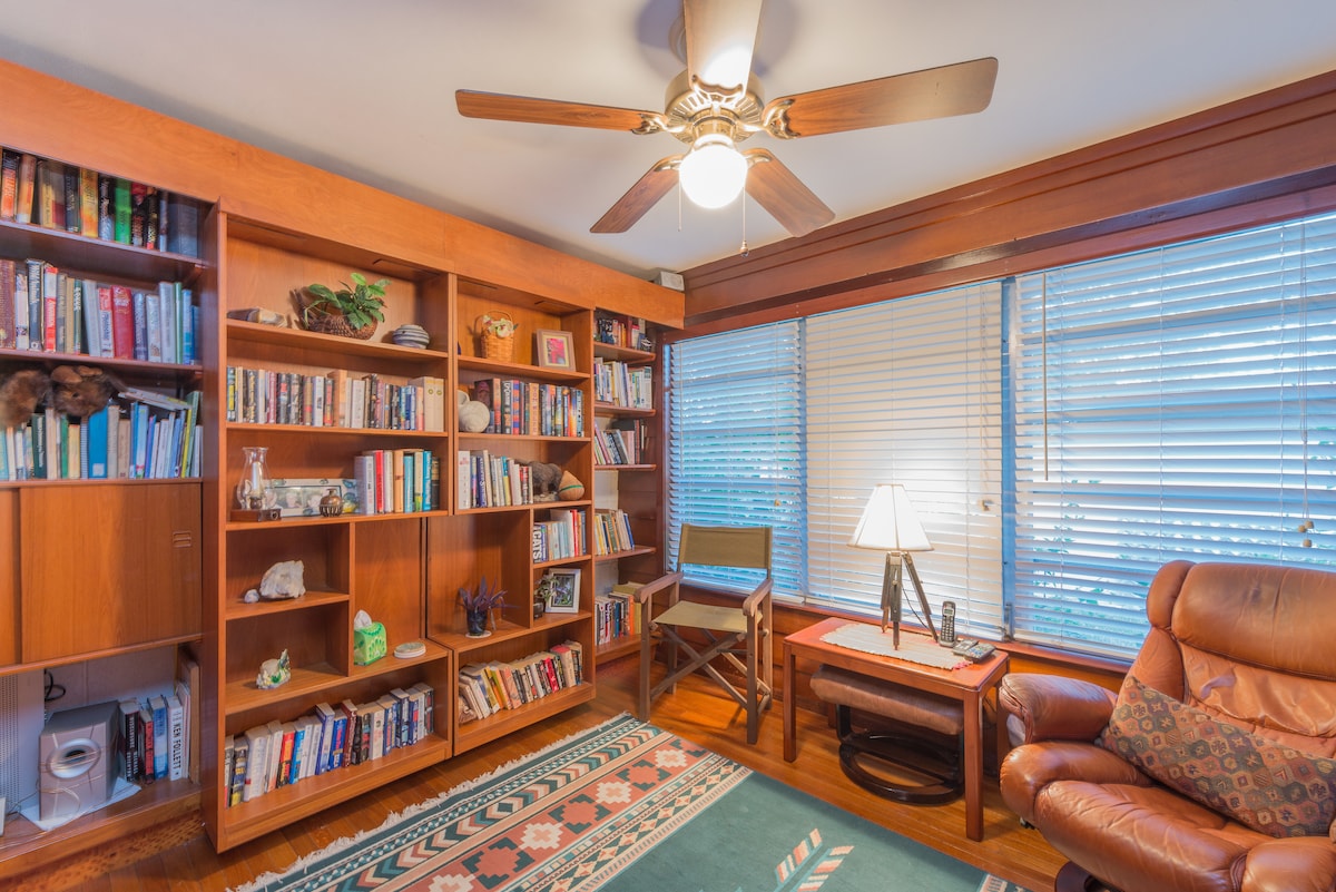 A cozy living area is displayed, featuring a wooden bookshelf filled with books and decorative items. Natural light filters through blinds on multiple windows. A comfortable leather chair and a small table with a lamp provide a welcoming reading space.