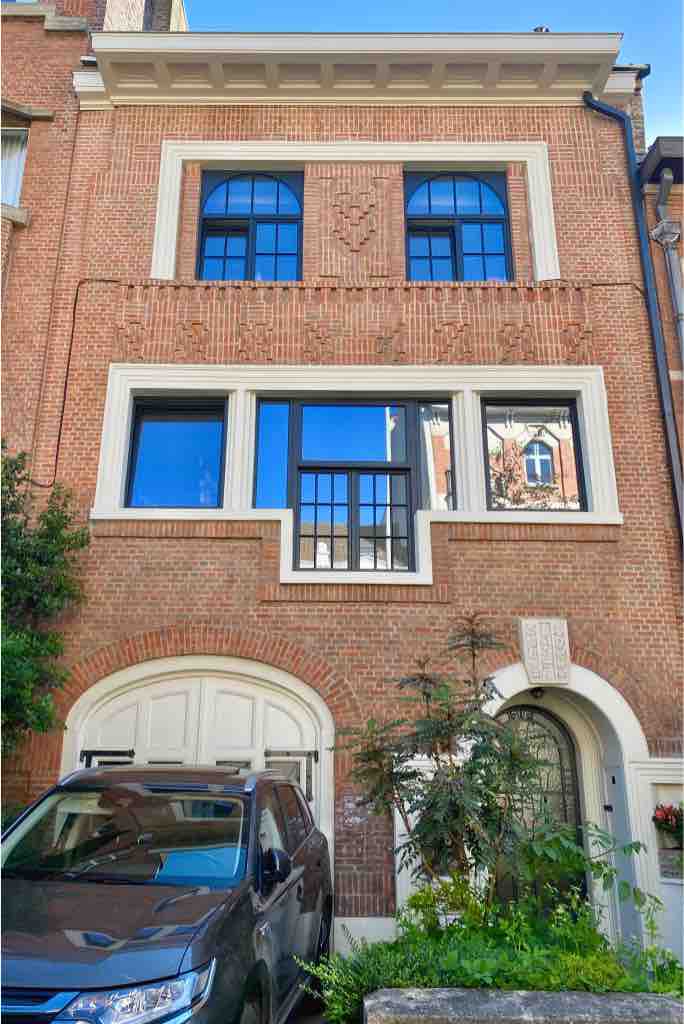 The exterior of the house features a red-brick facade with large windows that allow for ample natural light. Decorative details are noted above the entrance, and the garden area includes lush greenery. A vehicle is parked in front, enhancing the residential character.