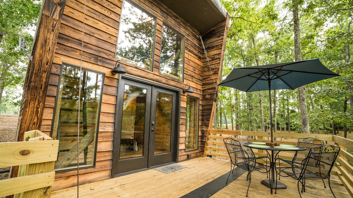 A charming treehouse exterior is showcased, featuring a combination of natural wood siding and large windows. A covered porch area is visible, furnished with a table and chairs, and an umbrella offers shade. Surrounding trees enhance the serene, woodland setting.