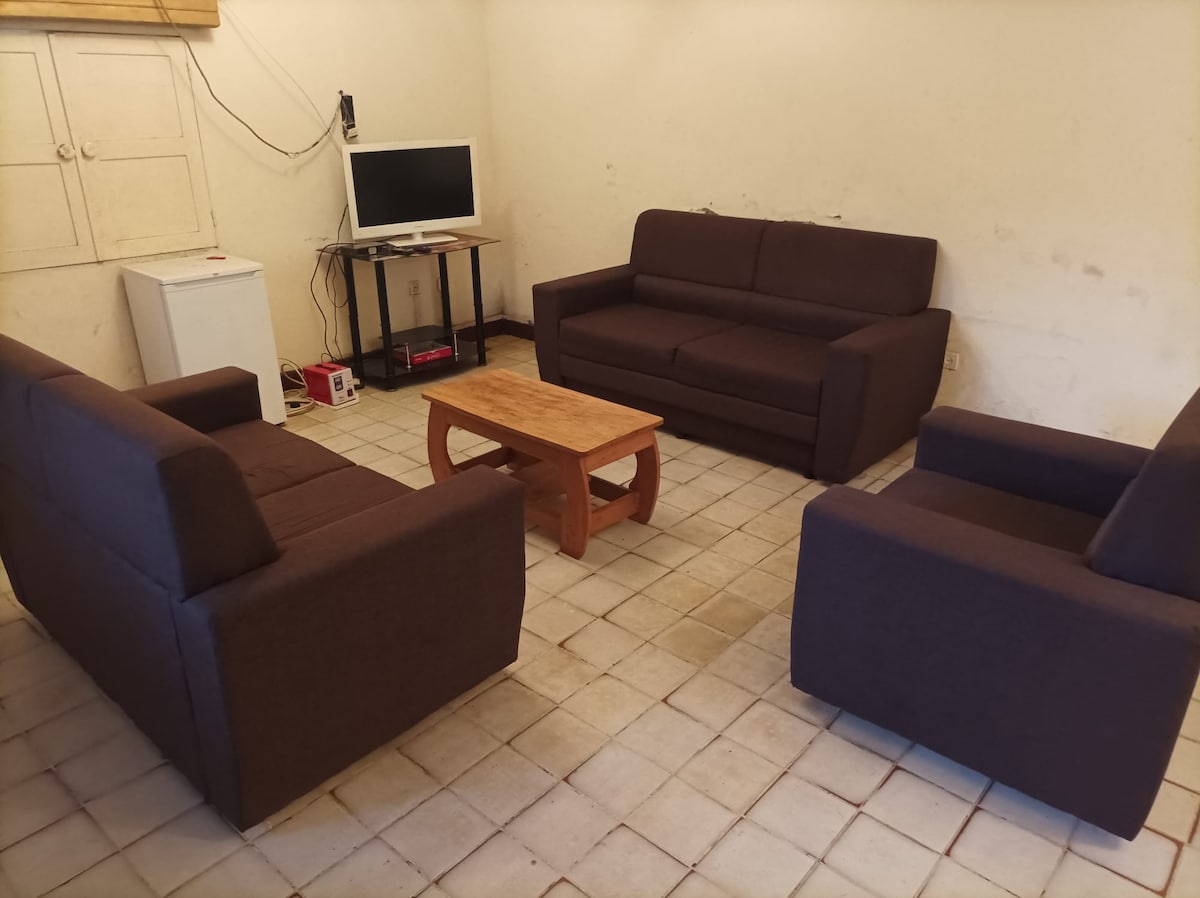 A living area features two sofas and an armchair arranged around a wooden coffee table. A small fridge is visible, along with a television mounted on the wall. The floor is tiled, and light enters through an unseen window.