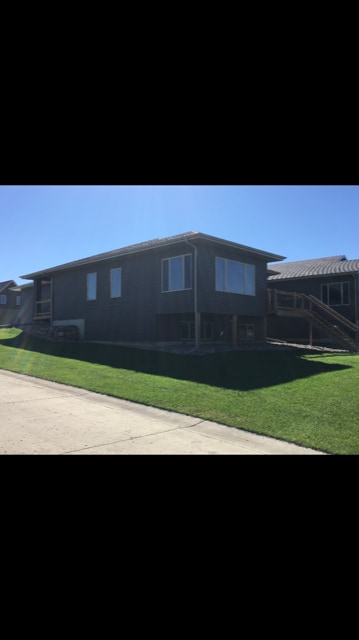 A modern cabin exterior is displayed, featuring a sloped lawn and a wooden deck with steps leading to the entrance. Large windows allow ample natural light, while the structured design complements a sunny day backdrop.