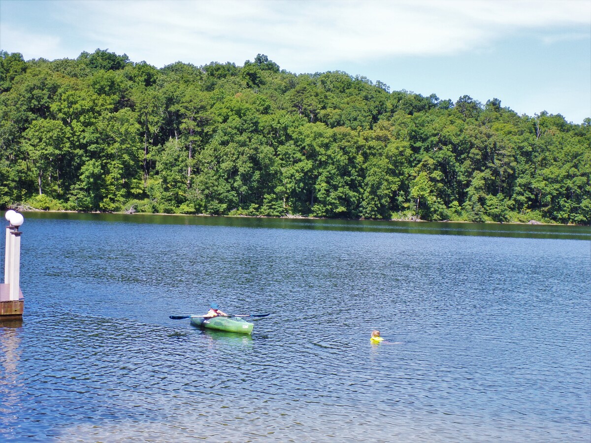 A serene lake scene features a person kayaking on calm waters, accompanied by a floating yellow duck. Lush green trees line the shore, reflecting in the water under a clear blue sky.