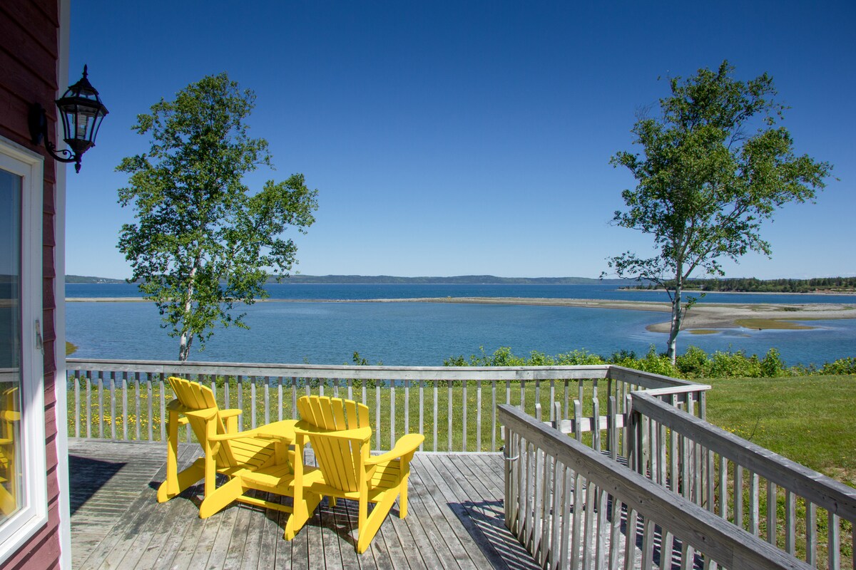 A wooden deck offers a clear view of the ocean and surrounding scenery. Two bright yellow Adirondack chairs are positioned for relaxation, while the bay's gentle tides and nearby trees create a tranquil setting. The expansive blue sky enhances the picturesque landscape.