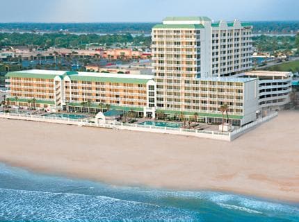 The beachfront resort is captured from an elevated perspective, highlighting its multi-story structure with green roofs and light-colored balconies. Pools can be seen in the foreground, surrounded by palm trees, with the sandy beach and ocean waves extending nearby.