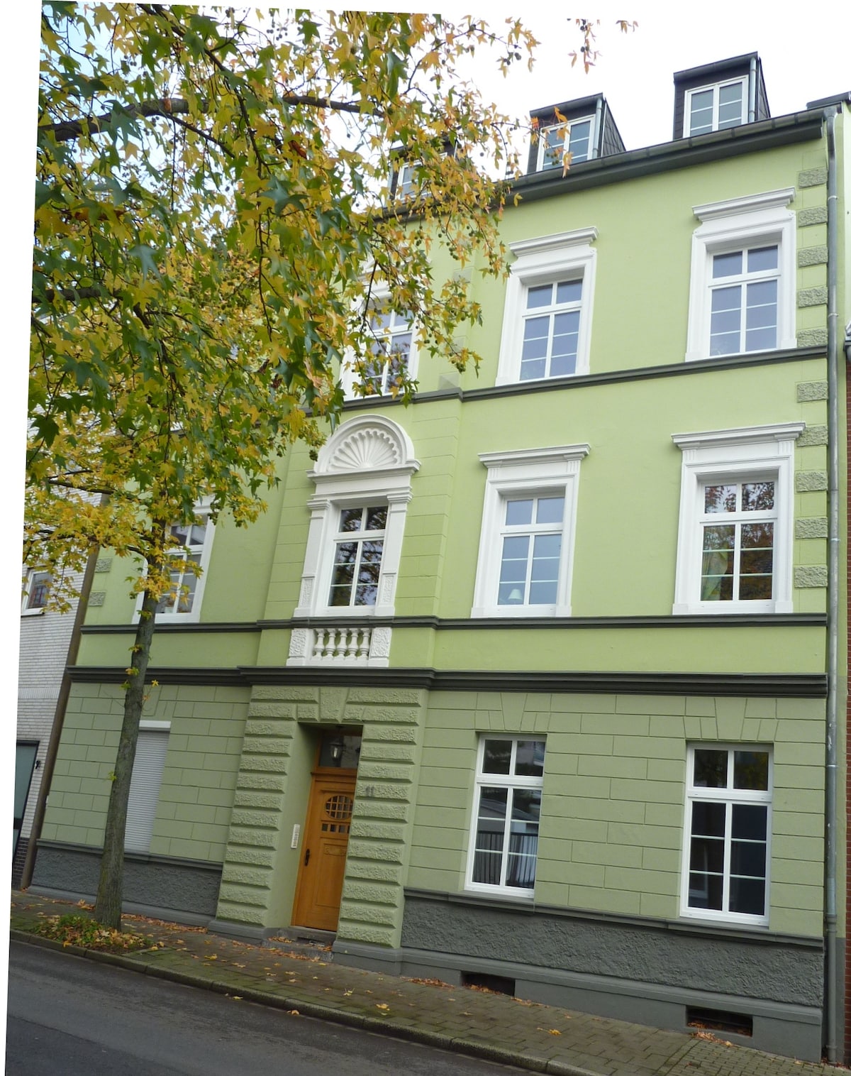 The exterior of a charming green building is displayed, featuring a decorative entryway and multiple windows with white trim. The structure is set among trees with autumn leaves, adding a touch of nature to the urban environment.