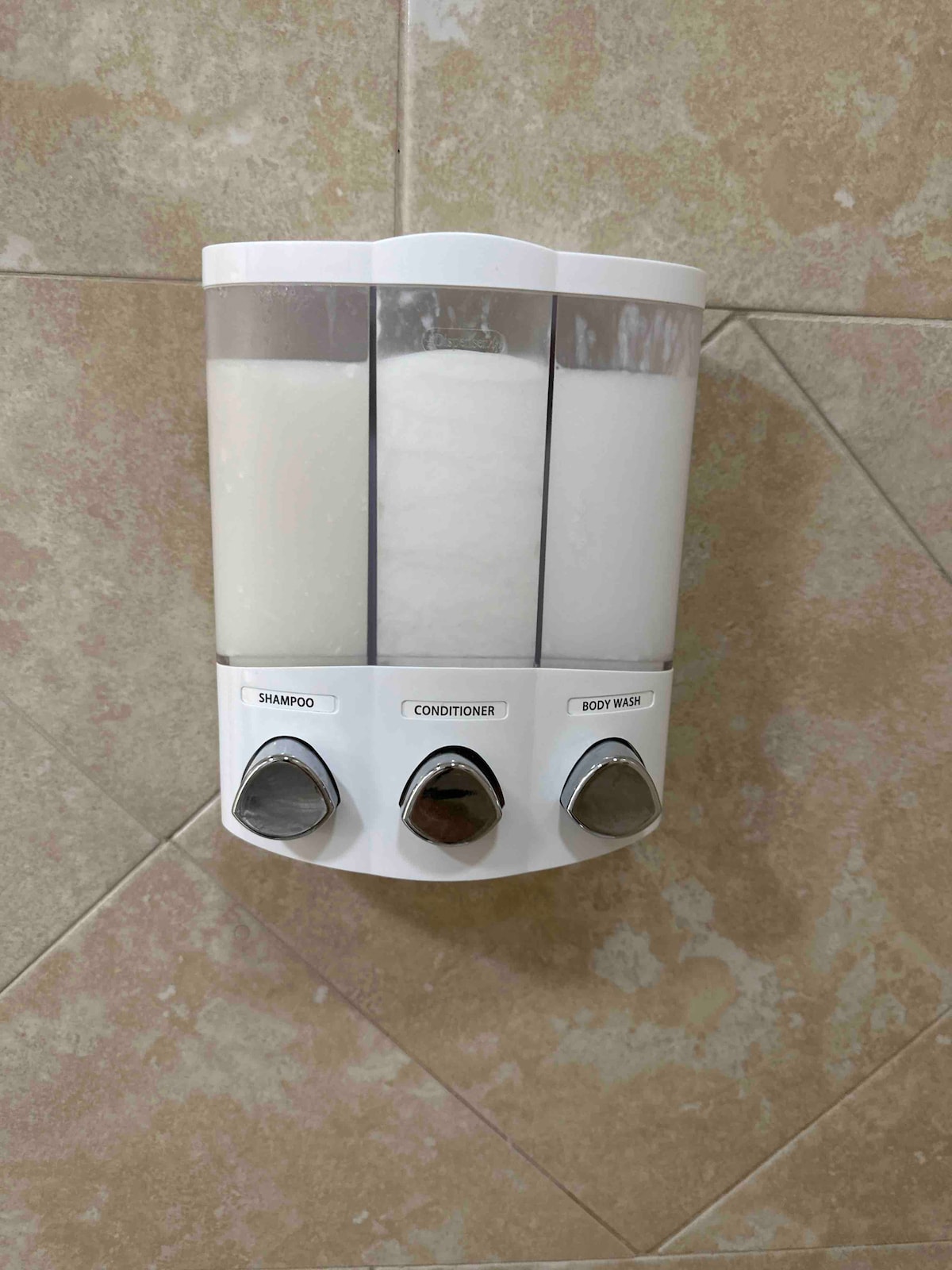 Three clear dispensers are mounted on the shower wall, labeled for shampoo, conditioner, and body wash. Each dispenser features a corresponding pump, designed for easy access during bathing. The background displays textured beige tiles, creating a clean and functional bathroom setting.