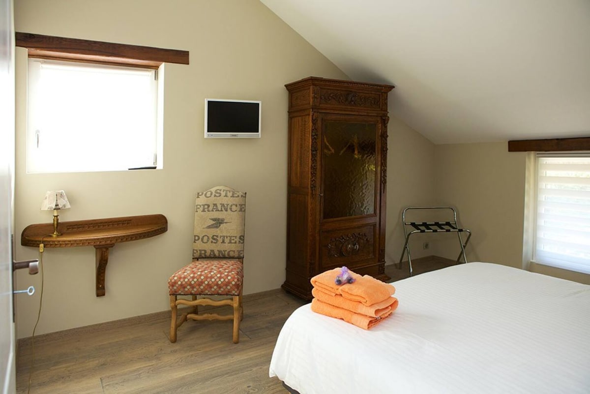 The sleeping area features a spacious bed with neatly arranged towels in orange. An antique armoire provides a touch of character, while a small wall-mounted television and a wooden shelf add functionality. Natural light enters through a window adorned with light shades.
