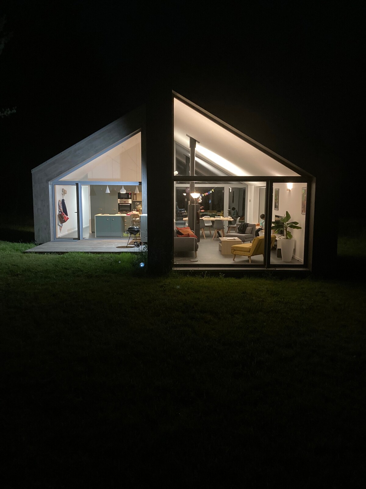 The modern wooden structure is illuminated from within, showcasing an open living space with a kitchen area and seating arrangements. Large glass doors provide a clear view of the cozy interior against the dark night landscape.