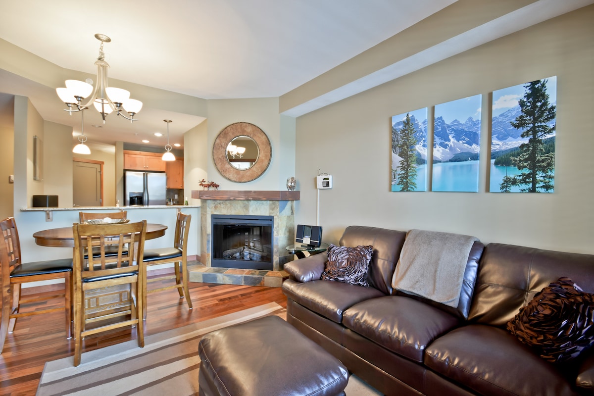 A welcoming living space features a leather sofa with decorative pillows and a cozy area rug. A dining table with wooden chairs is visible, and the room is highlighted by a fireplace and artwork showcasing a serene mountain landscape.