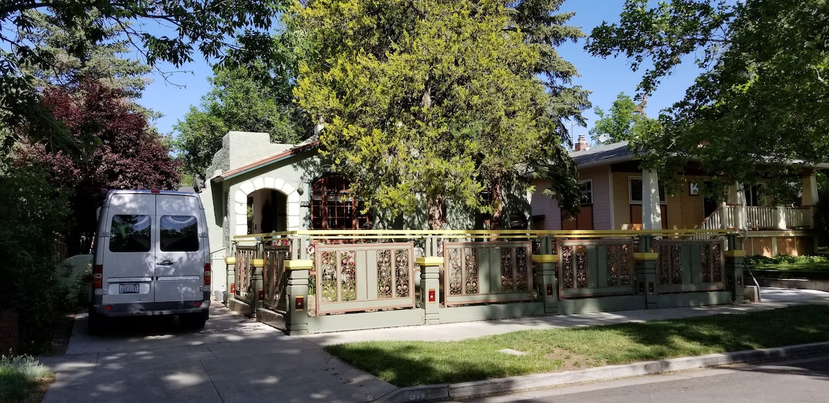 A quaint cottage exterior is surrounded by lush greenery, showcasing a decorative fence and a welcoming entryway. A sidewalk leads to the entrance, with a vehicle parked nearby. Large trees provide shade, enhancing the inviting character of the property.