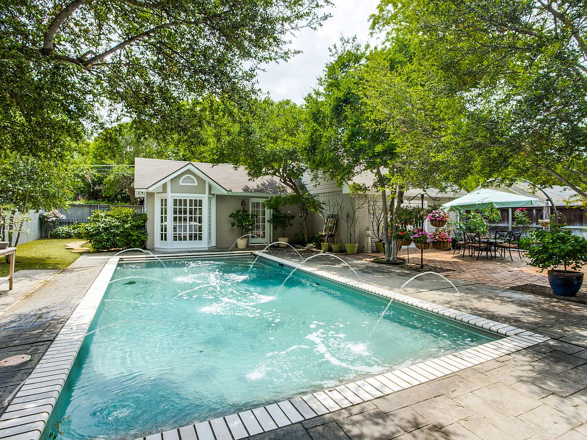 The image showcases a sparkling swimming pool surrounded by stone paving and lush greenery. Fountains add gentle water features, while shaded areas provide seating options. A neighboring house is visible in the background, indicating proximity to the guesthouse, enhancing the outdoor ambiance of the space.