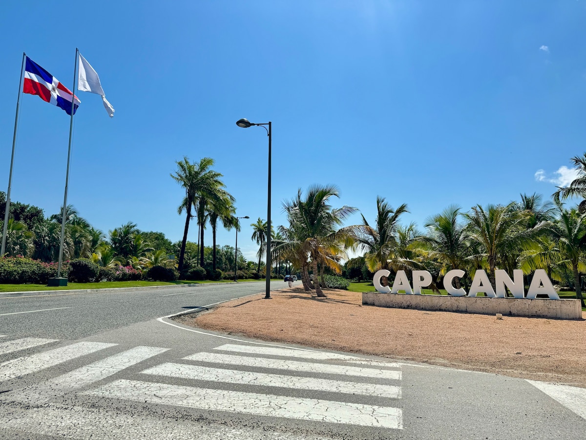 The entrance to Cap Cana is displayed with a large sign reading 'CAP CANA' set against a backdrop of landscaped gardens and palm trees. Clear blue skies and bright daylight emphasize the welcoming atmosphere, while national flags are visible fluttering gently in the breeze.