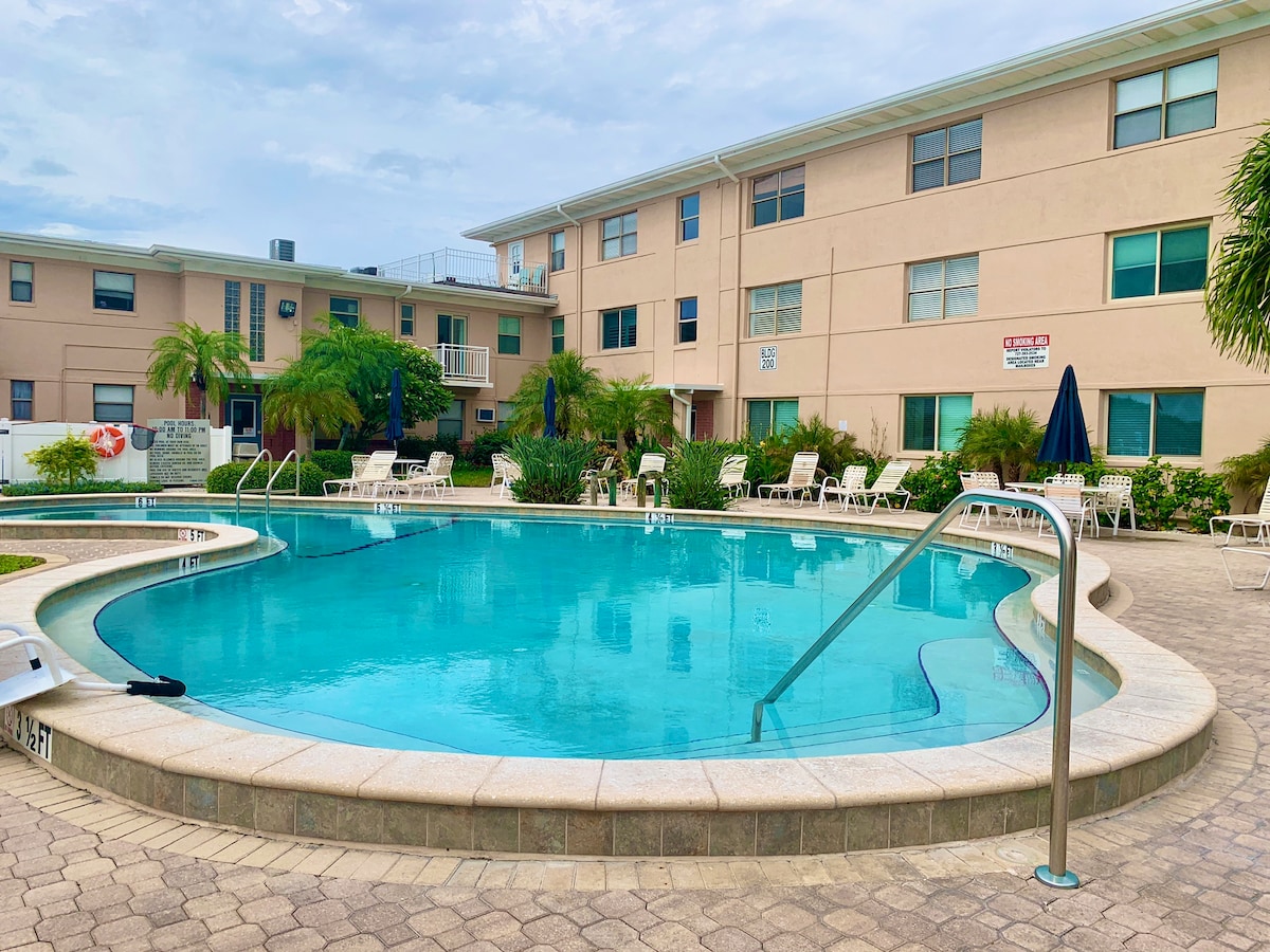 A resort-style pool is surrounded by lush greenery and palm trees, inviting relaxation. The water appears clear and inviting, with a gradual step leading in. Lounge chairs are arranged around the pool area, offering spaces for sunbathing. Nearby, a shaded umbrella stands for comfort.