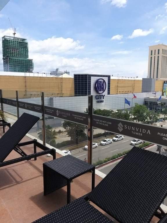 A balcony view from the 19th floor features modern seating overlooking SM City Cebu. The image captures the busy street below, with vehicles visible along the road. The surrounding area includes urban buildings and a clear sky, highlighting the city atmosphere.