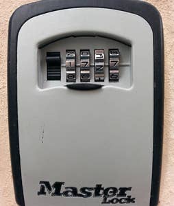 A wall-mounted lockbox is shown, featuring a combination dial and four numbered rotating dials. The box is designed for secure key storage, with the brand name 'Master Lock' visible at the bottom. The surface is a muted gray color.