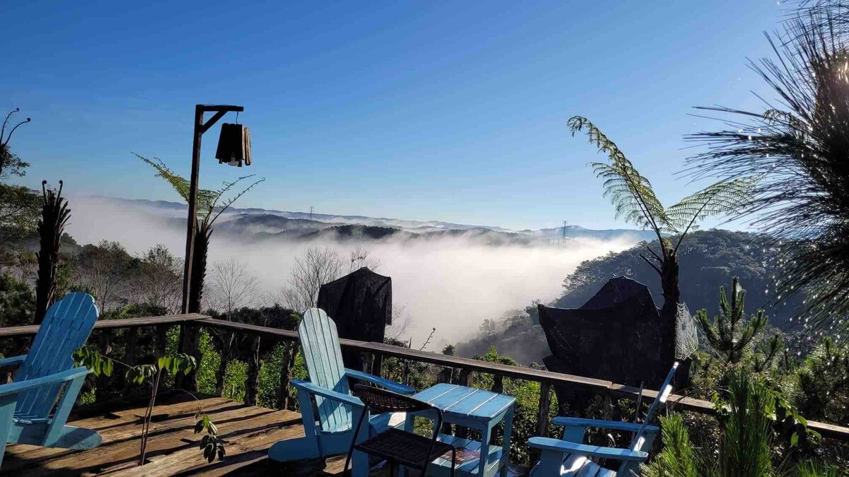 A deck features several blue Adirondack chairs, positioned to overlook a misty valley. Lush greenery and trees frame the scene, while fog blankets the landscape below, creating a serene backdrop. A small lantern hangs nearby, adding a gentle touch to the outdoor space.