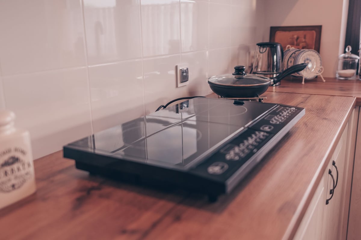 A modern kitchen counter showcases a sleek induction cooktop with a black surface, complemented by a frying pan and a kettle. Natural light filters in, reflecting off the glossy surface, while the warm wood of the countertop adds an inviting contrast.