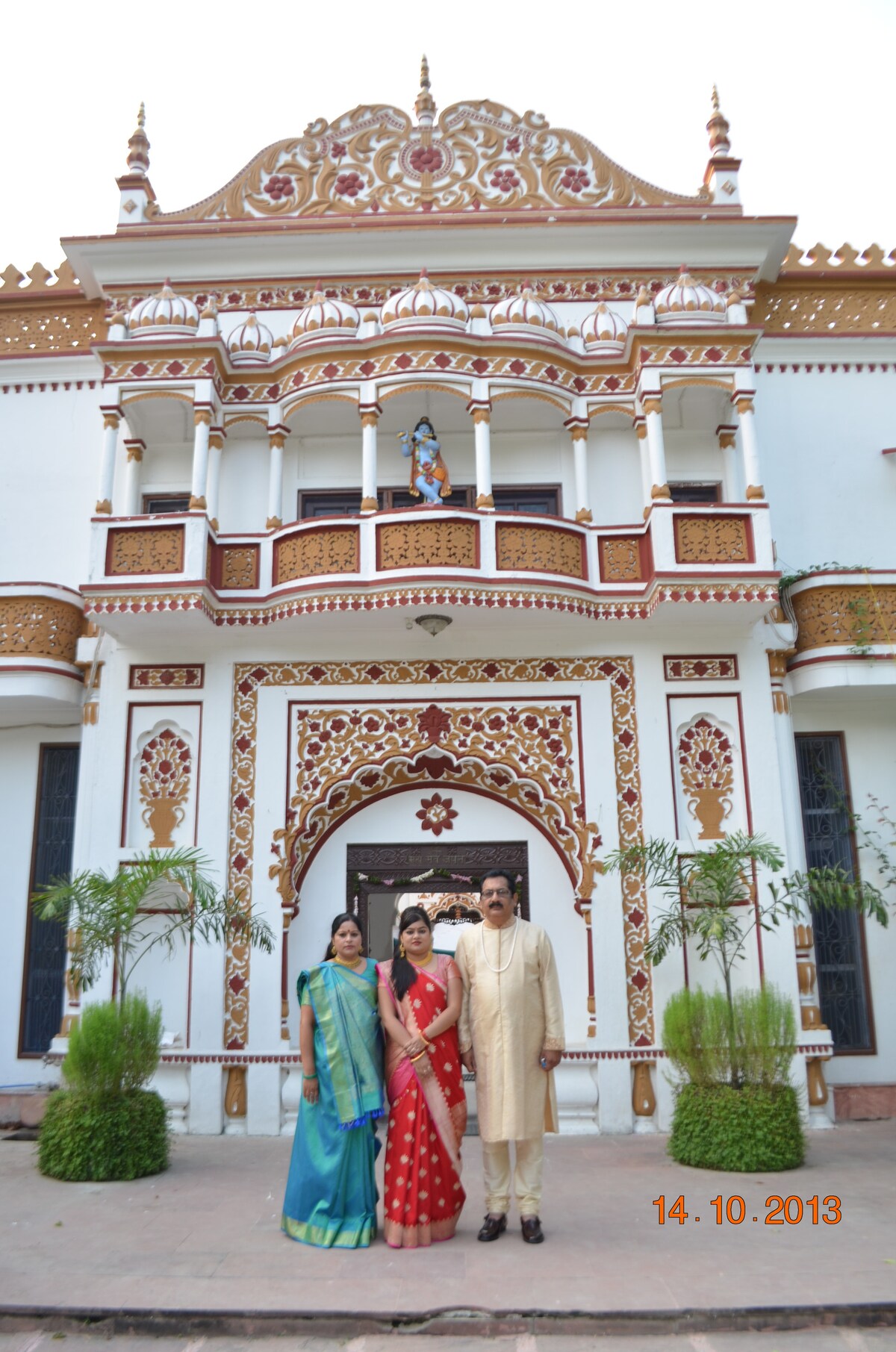 An intricately designed building serves as the backdrop, featuring ornate architectural details and vibrant decorations. Flanking the entrance are well-maintained plants, adding a touch of greenery to the exterior. The elegant facade highlights cultural craftsmanship and invites visitors to appreciate its unique style.