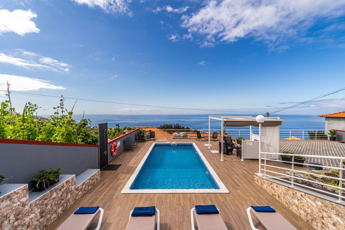 An outdoor space features a rectangular swimming pool surrounded by sun loungers. Lush greenery is visible along the sides, while ocean views extend in the background under a clear sky. A covered seating area can be seen at one corner, offering relaxation options.