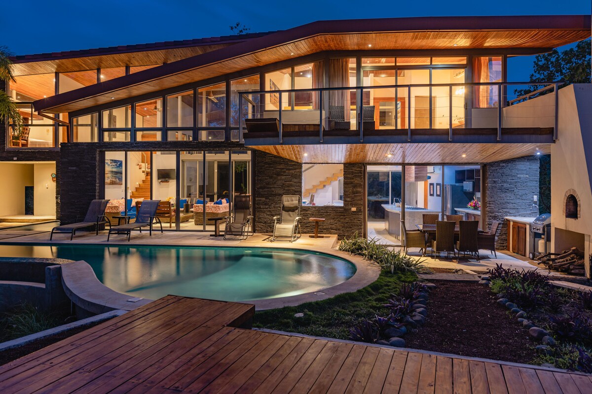 The modern house is illuminated at night, showcasing its architectural lines and large glass windows. The infinity-edge pool reflects the warm lights, with lounge chairs positioned nearby. An outdoor seating area is visible, inviting relaxation around the pool and garden landscape.