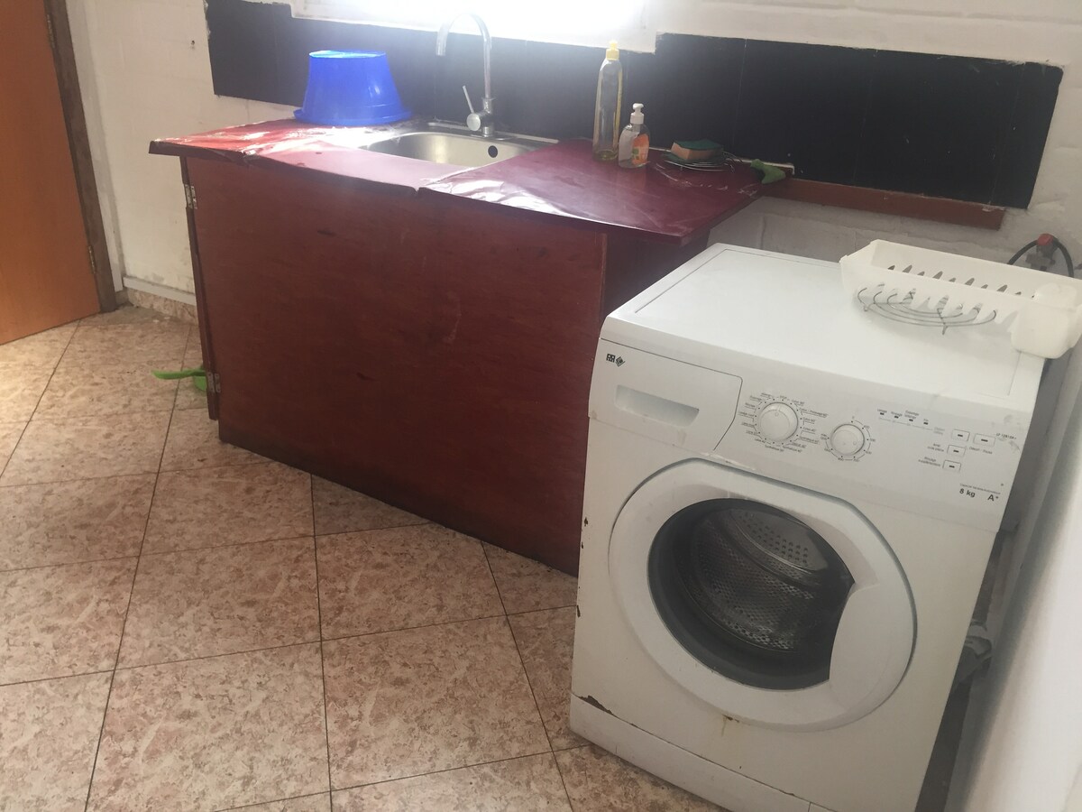 A compact kitchen area features a wooden countertop with a small sink. A washing machine is positioned next to the countertop. Tile flooring is visible, along with a blue basin and various cleaning supplies on the counter.