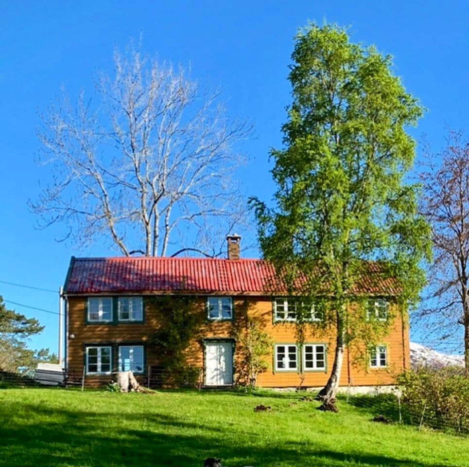 The two-story wooden farmhouse boasts a vibrant red roof and multiple white-framed windows. Surrounding greenery highlights the house's traditional architecture, and tall trees provide a natural backdrop on a clear blue sky day.