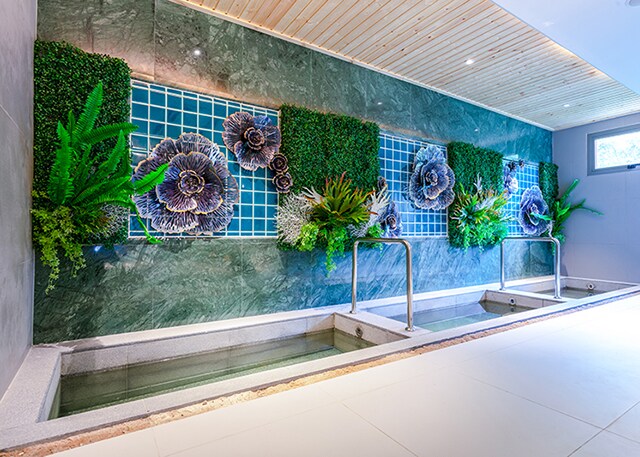A serene spa area features a stone wall adorned with artistic floral decorations, surrounded by greenery. Two shallow pools are present, inviting relaxation. Soft lighting enhances the tranquil ambiance, designed to promote a calming atmosphere.