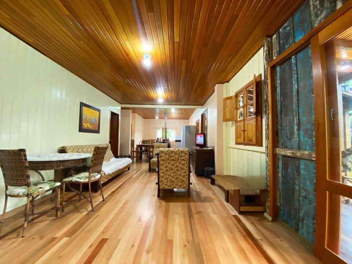 A spacious living area is showcased, featuring wooden flooring and a wooden ceiling. A dining table is present alongside wicker chairs, with a cozy couch and TV in view. Natural light filters through windows, creating a warm and inviting environment.