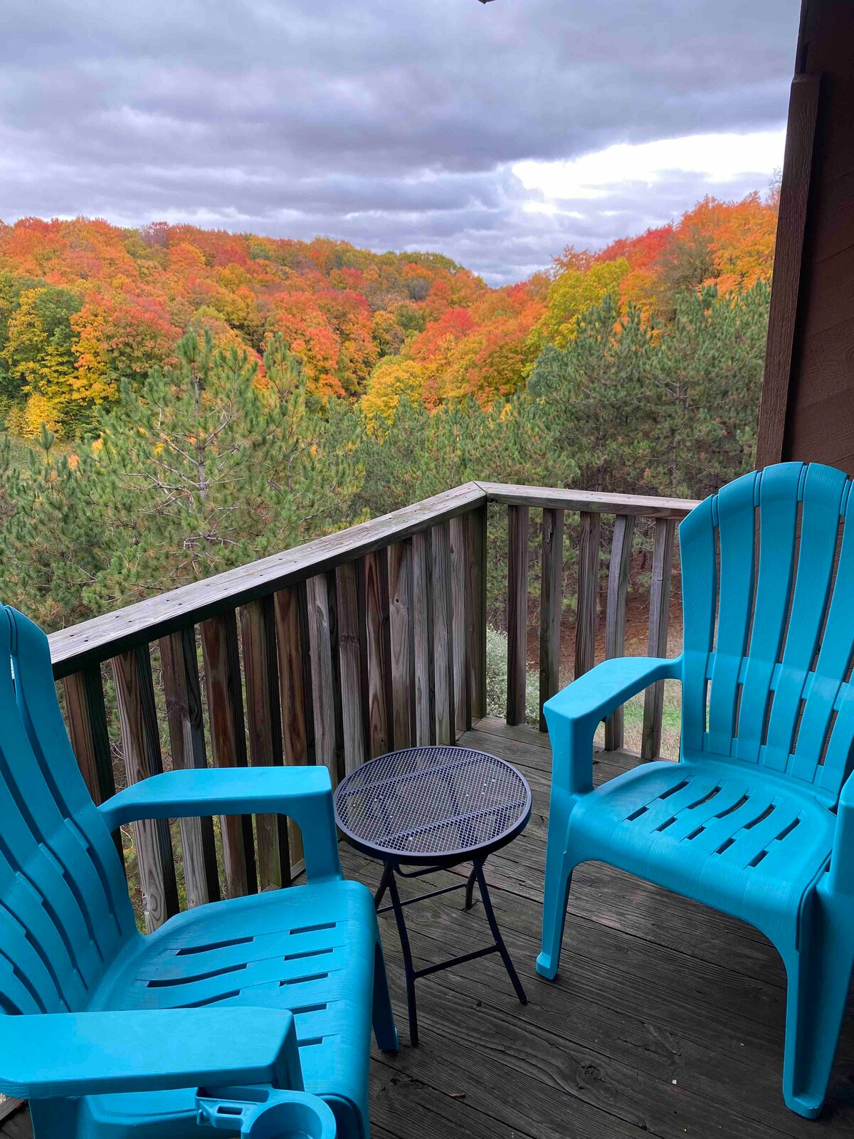 A private deck features two bright blue chairs and a small black table, set against a backdrop of vibrant autumn foliage. The trees display hues of orange, yellow, and green, while a cloudy sky adds to the serene atmosphere.