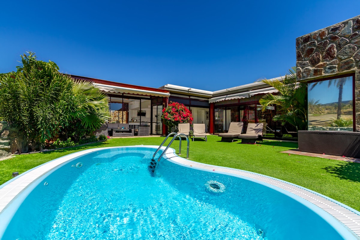 A private heated swimming pool with clear blue water is framed by lush greenery and landscaped grass. Sun loungers are arranged nearby, inviting relaxation under the clear blue sky. The villa's modern exterior and large windows create a harmonious connection with the garden.