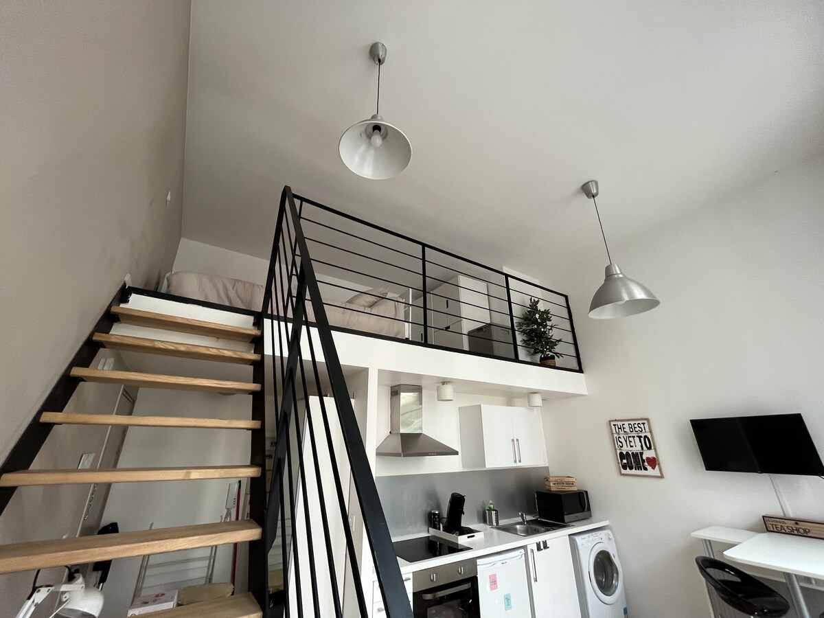 A spacious loft-style apartment with a wooden staircase leading to a mezzanine sleeping area. The kitchen is equipped with modern appliances and a washing machine, while pendant lights hang from the ceiling. A small potted plant adds a touch of greenery to the decor.
