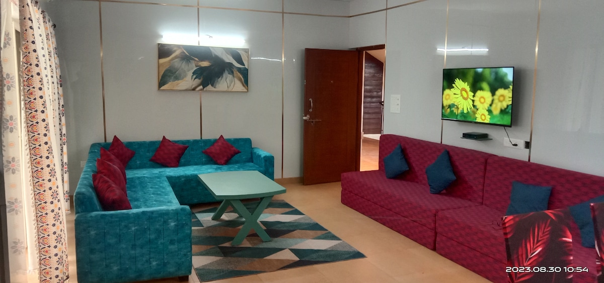 The living room features a vibrant arrangement of two large sofa sections in teal and burgundy, accented with colorful cushions. A light green coffee table sits at the center on a patterned rug. A wall-mounted flat-screen TV and a piece of artwork complete the inviting space.
