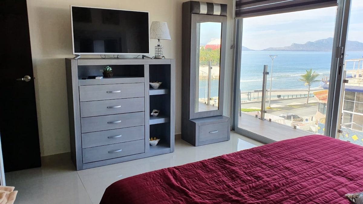 A modern bedroom features a spacious bed with a deep red blanket, a sleek dresser with multiple drawers, and a large flat-screen television mounted above. A mirror reflects light from the expansive windows that showcase a view of the ocean and coastal landscape.