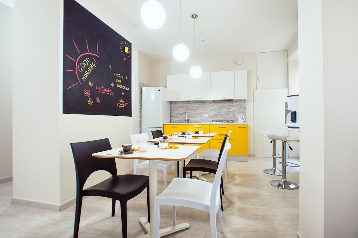A modern kitchen space features a bright yellow cabinetry, complemented by a clean white dining table with black and white chairs. An inviting atmosphere is created with overhead pendant lights illuminating the area. A chalkboard wall adds a playful touch to the design.