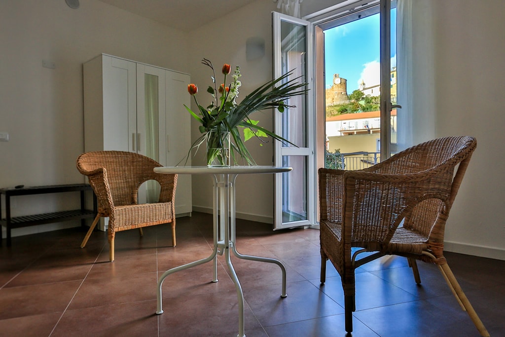 A bright seating area is presented, featuring two woven chairs positioned around a small glass table. A vase of fresh flowers adds a touch of color. Natural light floods the space through a partially open window, which offers a view of the surrounding landscape.