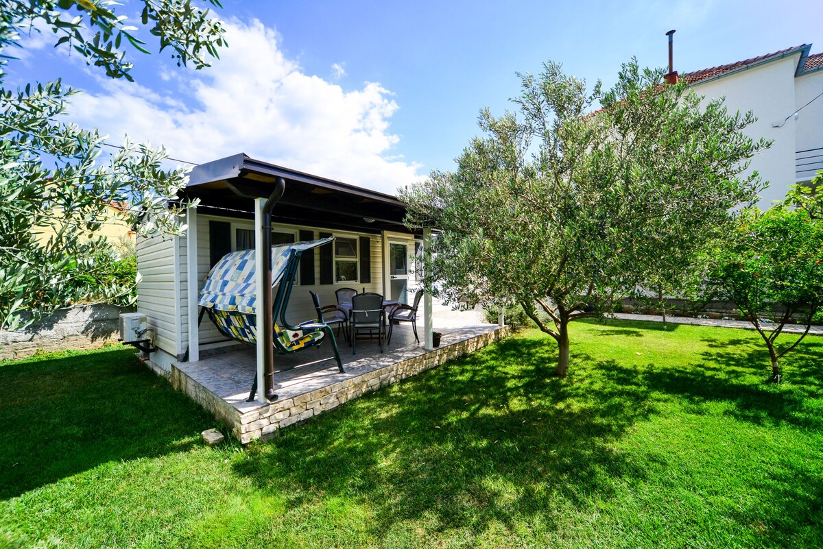 An outdoor seating area is shown, featuring a covered patio with colorful hammocks and several chairs. A grassy area surrounds the patio, complemented by a tree providing additional shade. The backdrop includes a residential structure and bright blue sky with scattered clouds.